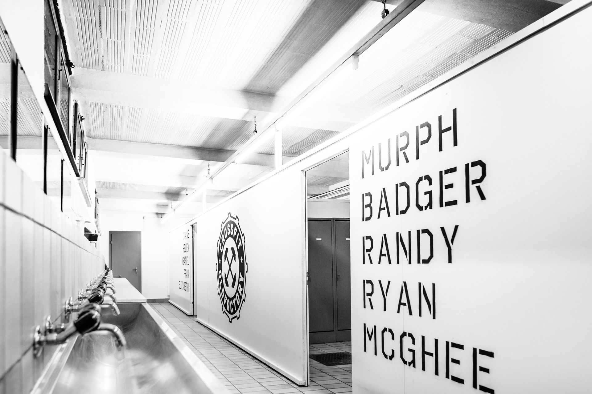 Schwarz-Weiß-Foto einer Umkleidekabine. Namen sind auf die Türen gedruckt, Waschbecken sind im Vordergrund zu sehen.