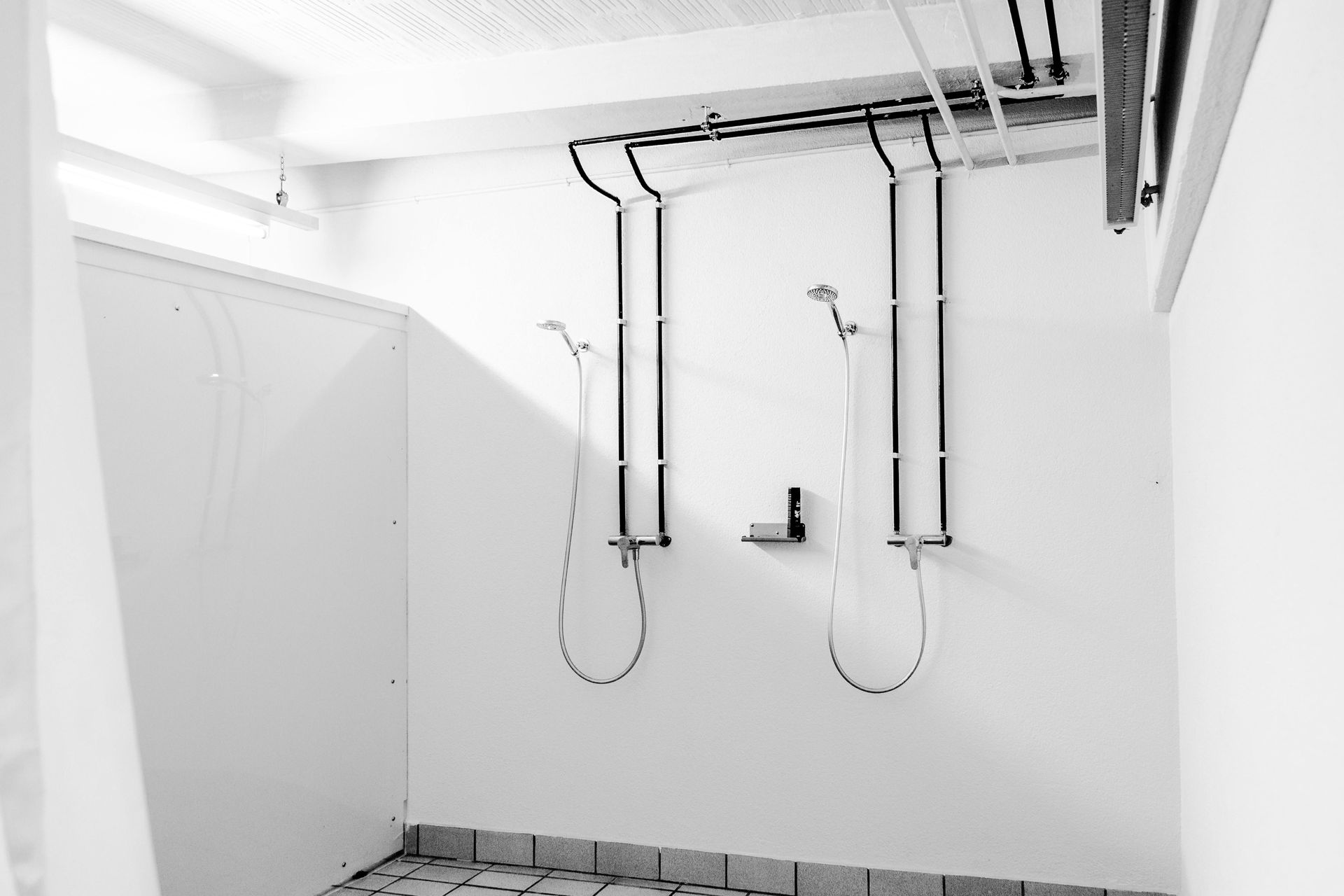 Zwei öffentliche Duschkabinen mit darüberliegenden Rohren und Duschköpfen vor einer weißen Wand.