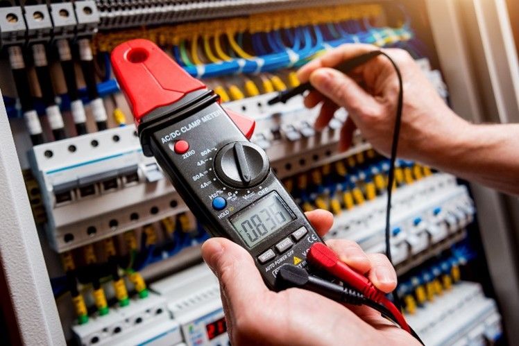 A person is holding a digital multimeter in front of a electrical cabinet.