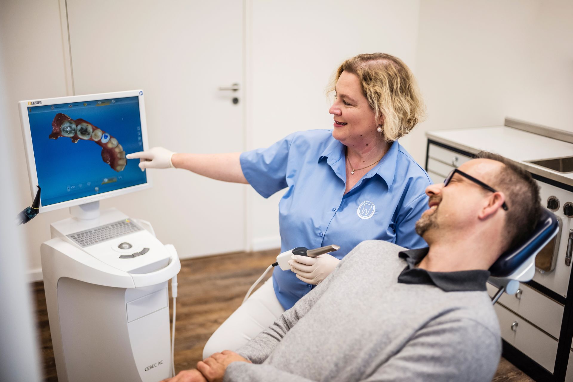 Eine Frau zeigt auf einen Computerbildschirm, während ein Mann auf einem Zahnarztstuhl sitzt.