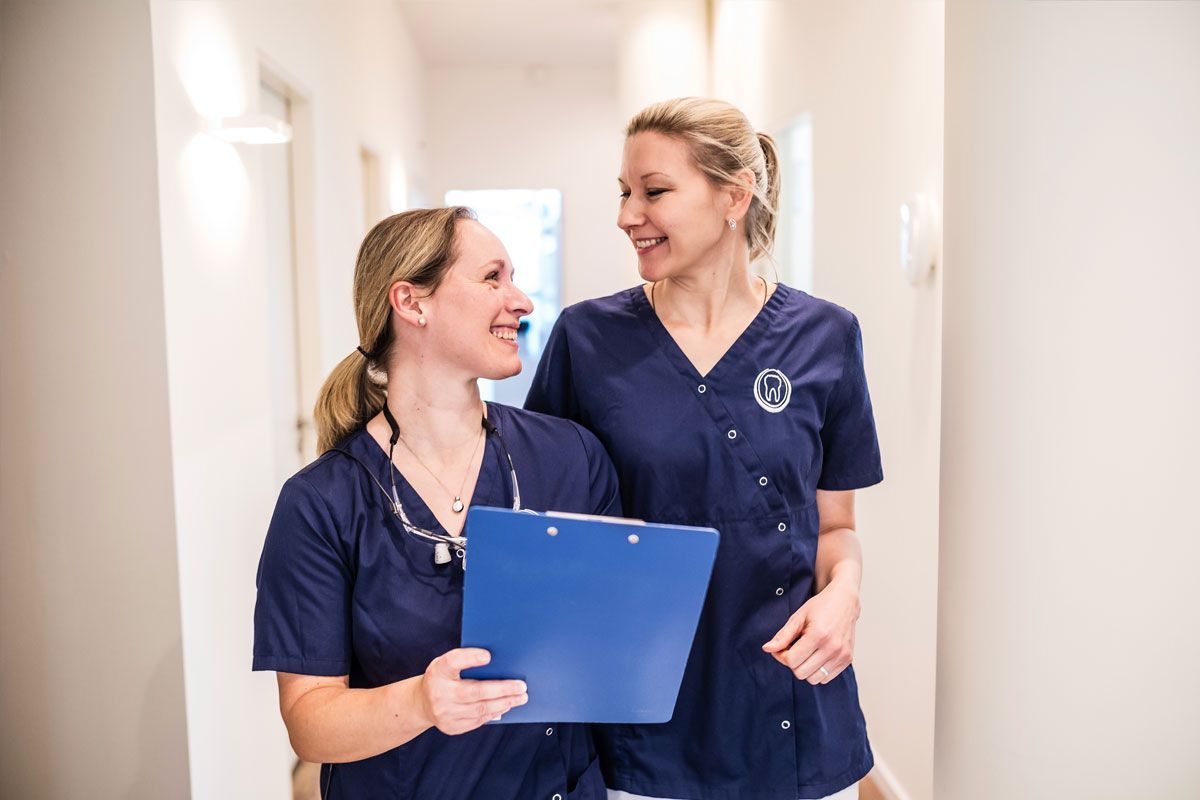Zwei Krankenschwestern stehen mit einem Klemmbrett in der Hand nebeneinander in einem Flur.