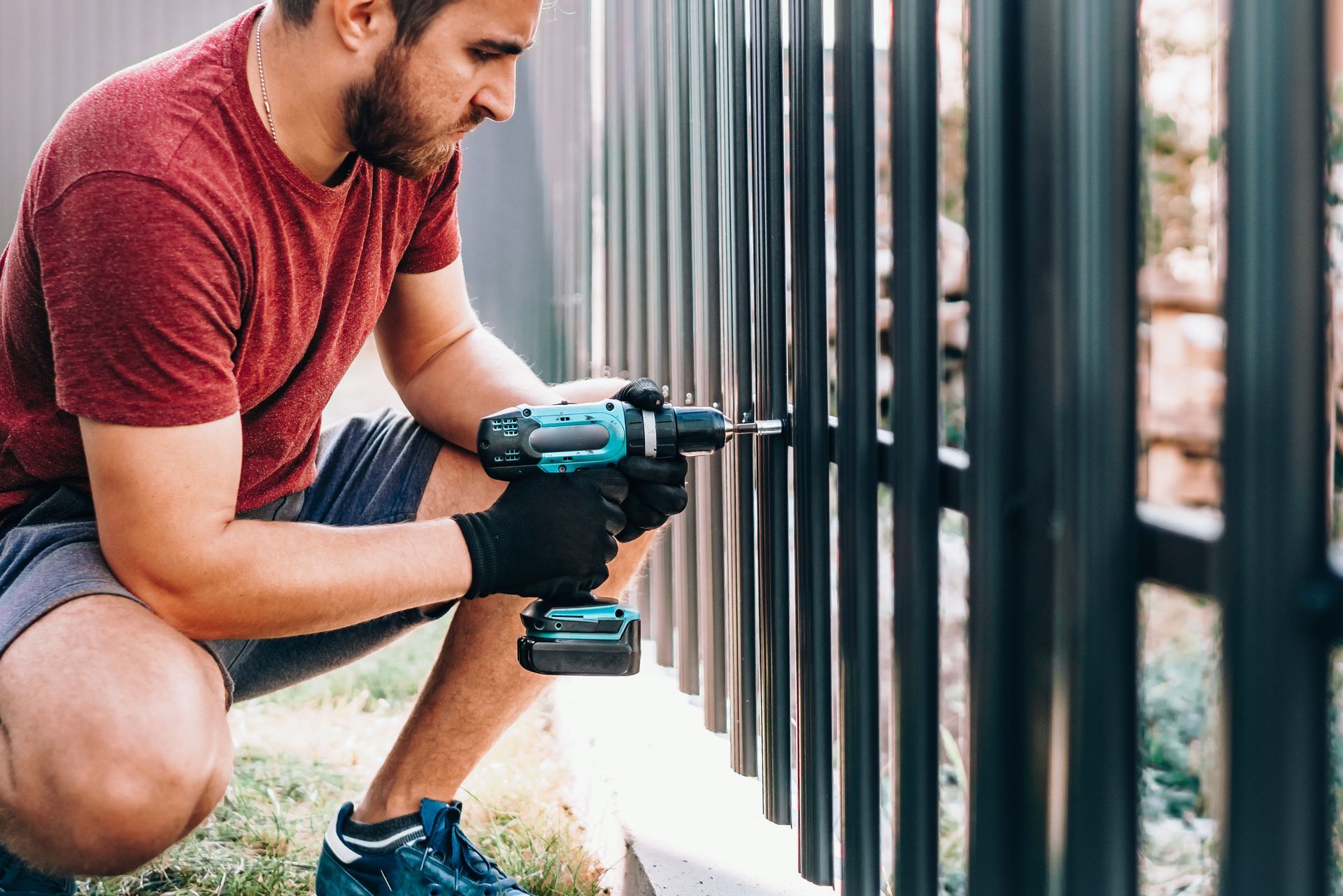 Man in red shirt and shorts using a drill to install a black metal fence outdoors