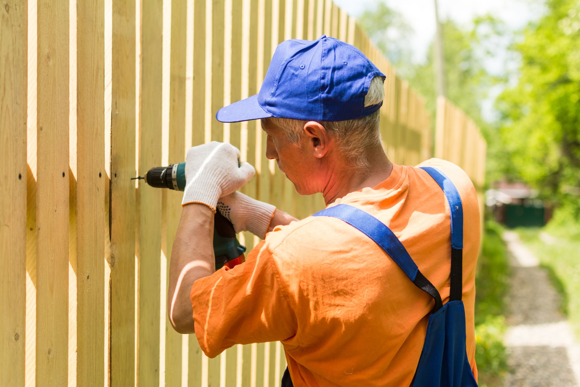 Man in orange shirt and blue cap uses a drill on a wooden fence outdoors