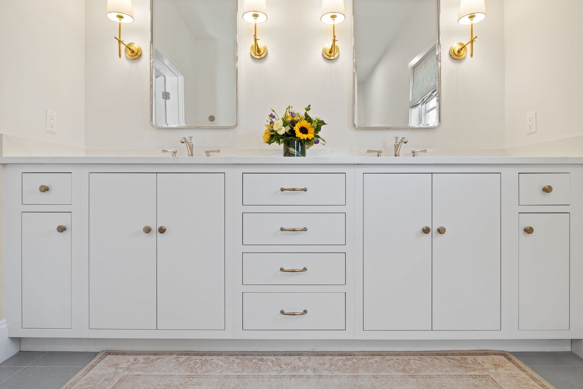 White bathroom vanity with double sinks, mirrors, brass sconces, and a sunflower vase on the counter