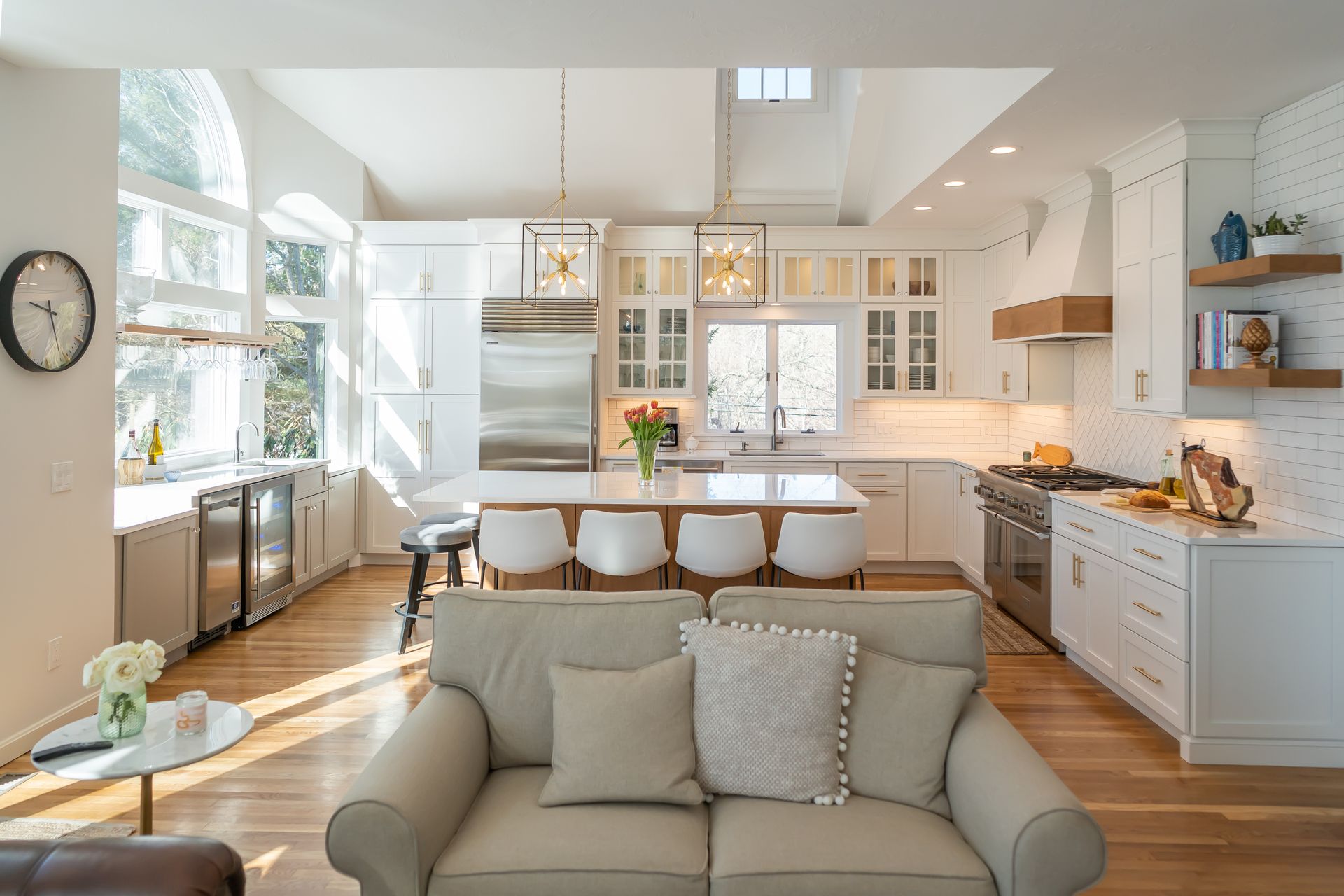 Bright modern kitchen and living area with white cabinets, island stools, and sunlit sofa in the foreground