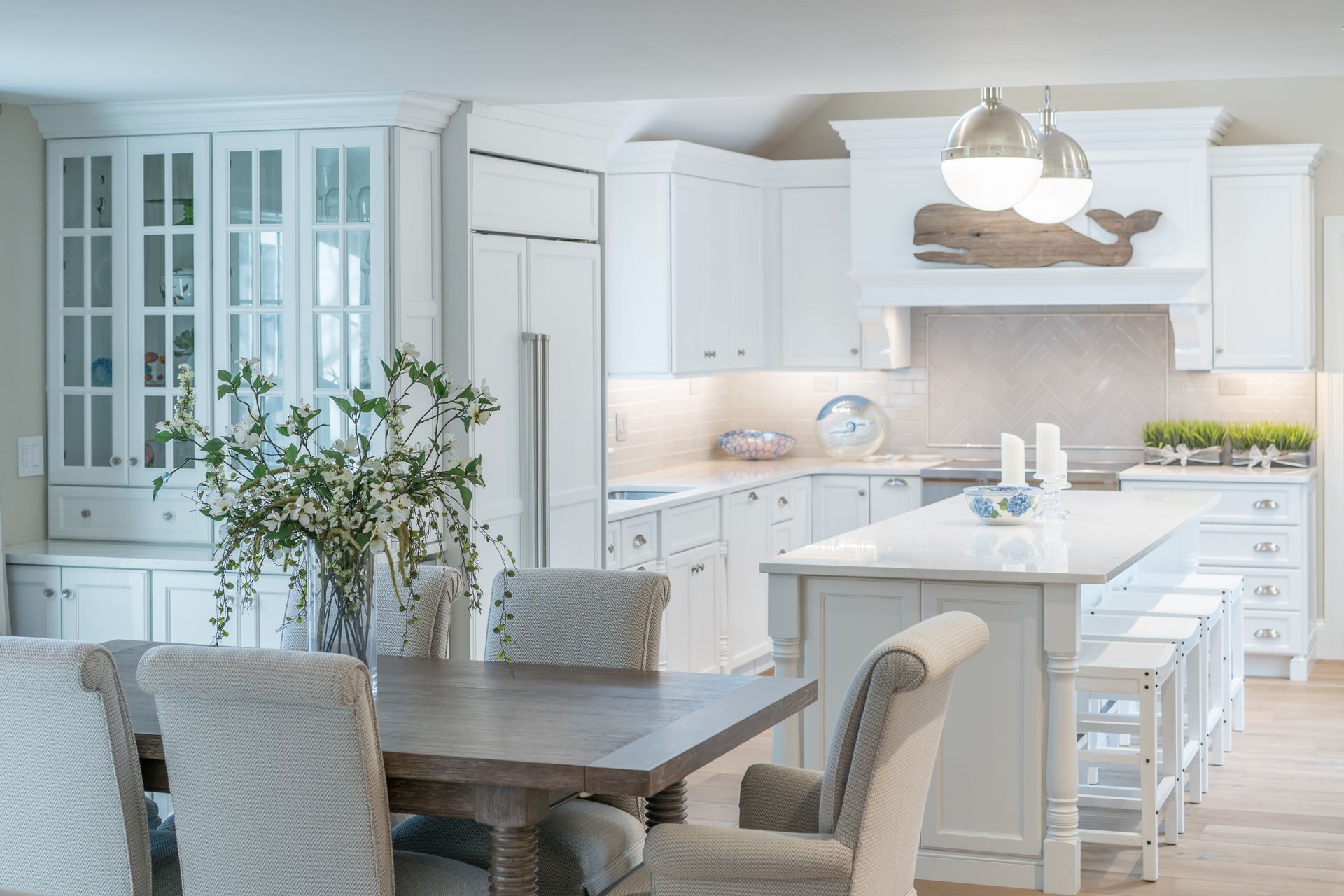 Bright white kitchen and dining area with island, pendant light, glass cabinets, and wooden table with chairs