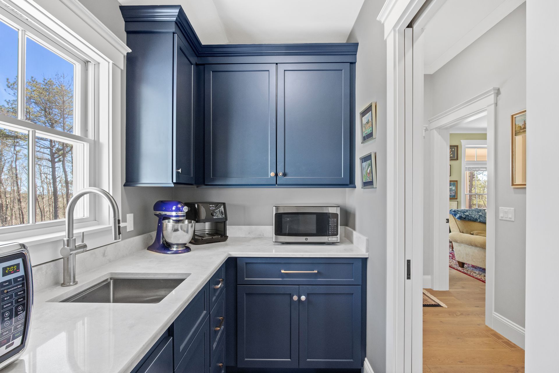 Compact kitchen with navy cabinets, white countertops, sink, microwave, and a view into a bright adjoining room