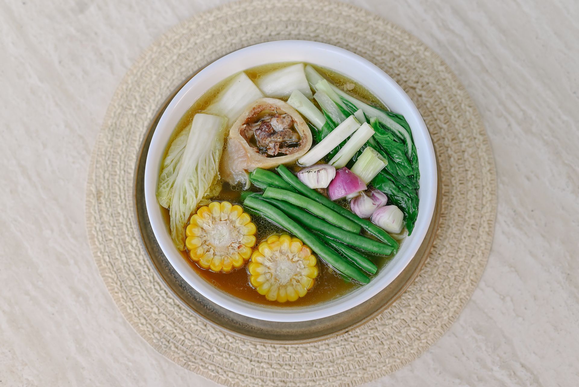 A bowl of soup with corn , green beans , onions and cabbage on a table.
