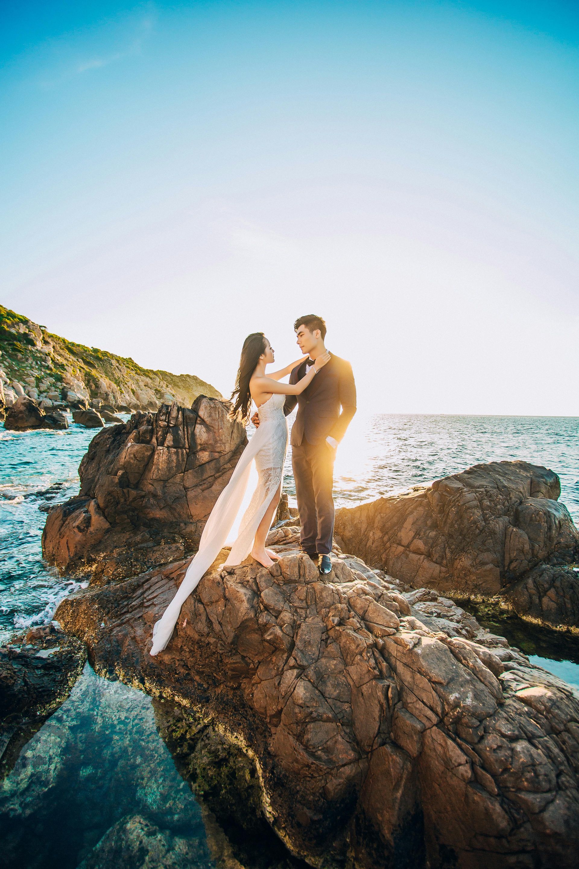A bride and groom are standing on a rock near the ocean.