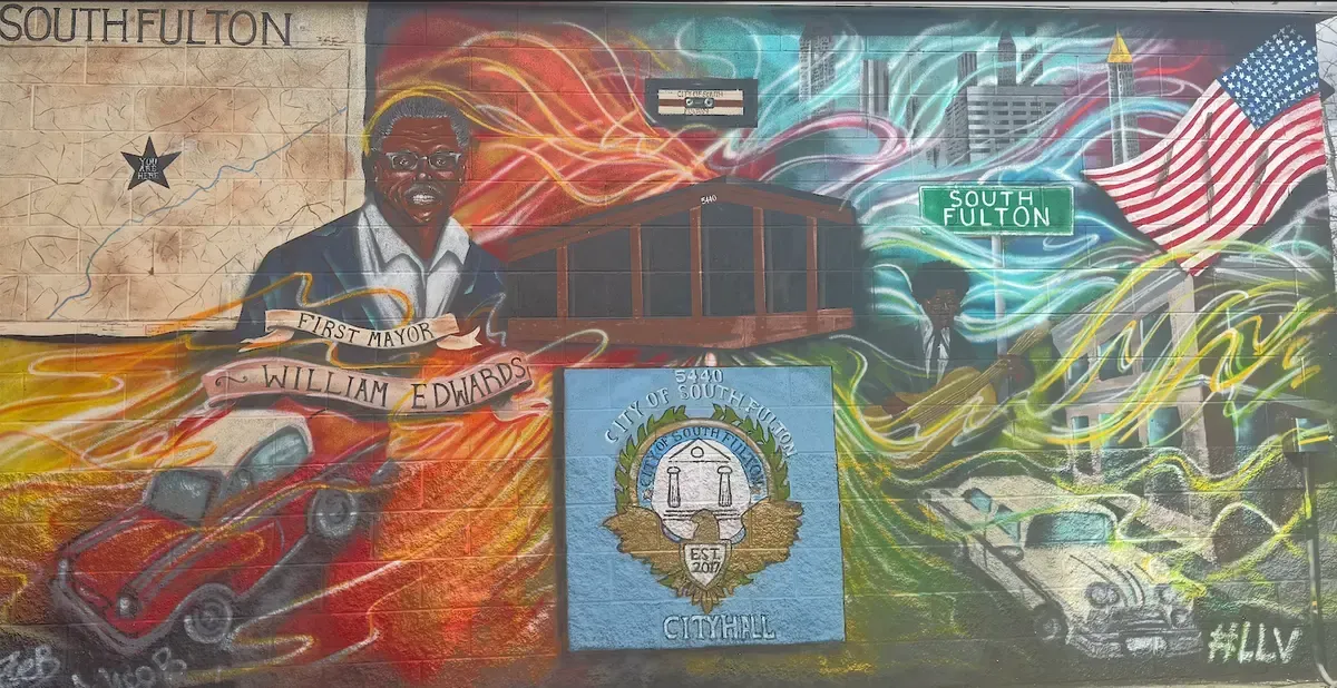 Mural in South Fulton: portraits, cars, flag, city with energetic color scheme, reflecting community and history.