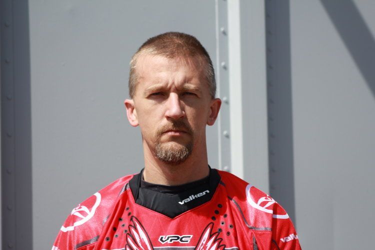 A man with a beard and a red shirt is standing in front of a wall.