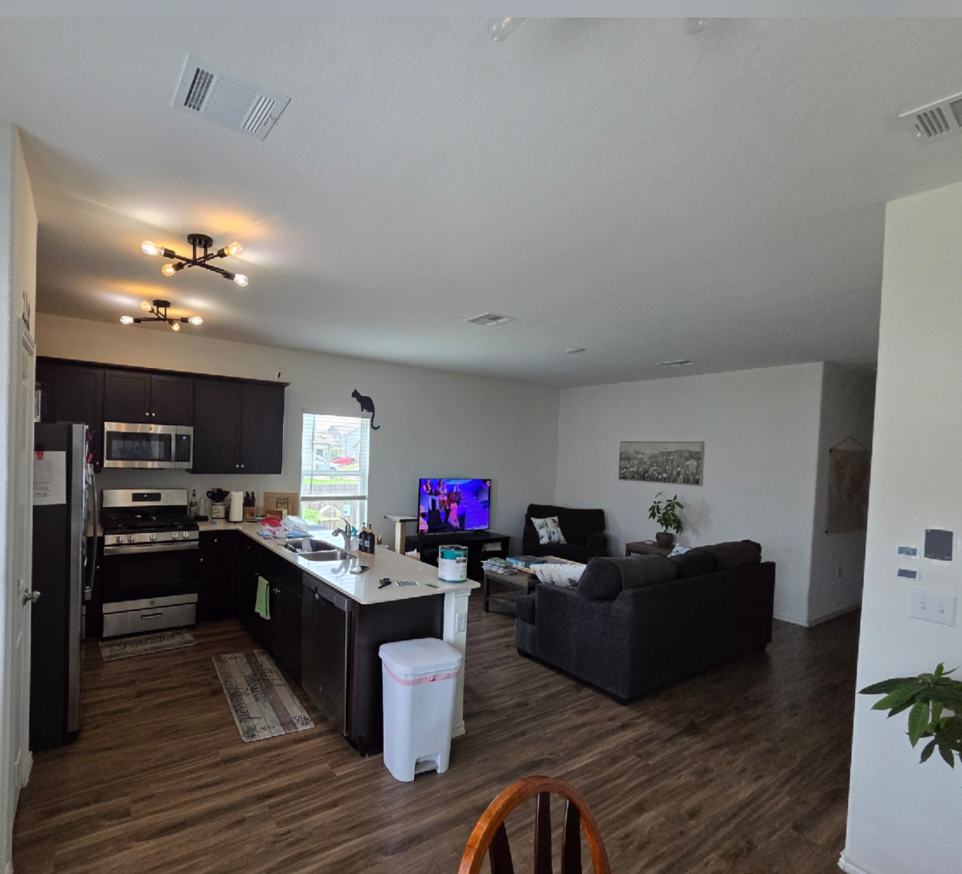 Open kitchen and living room with dark cabinets, wood floors, gray sofa, and TV on a stand