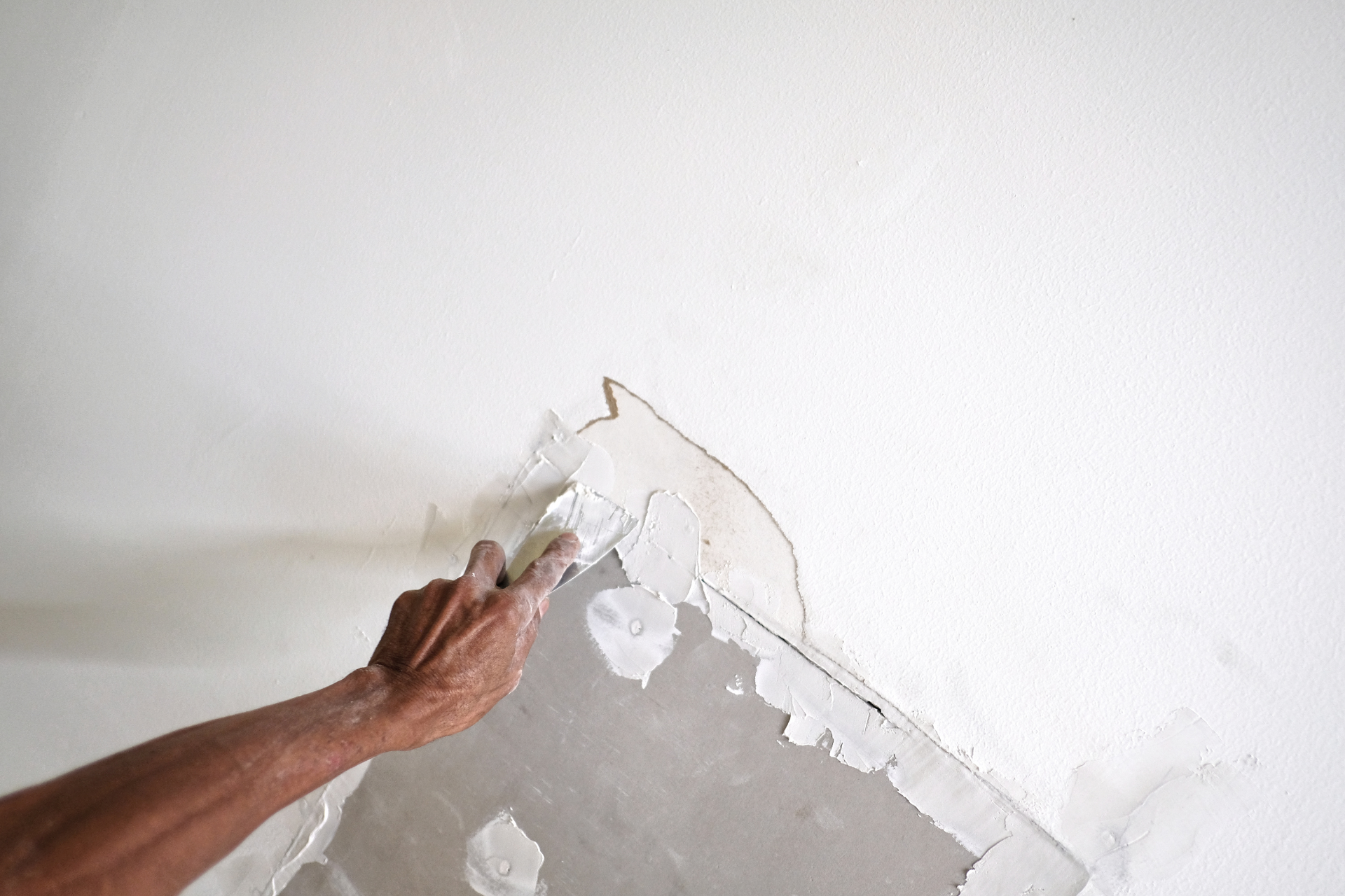 Hand scraping peeling white paint from a wall corner over gray plaster