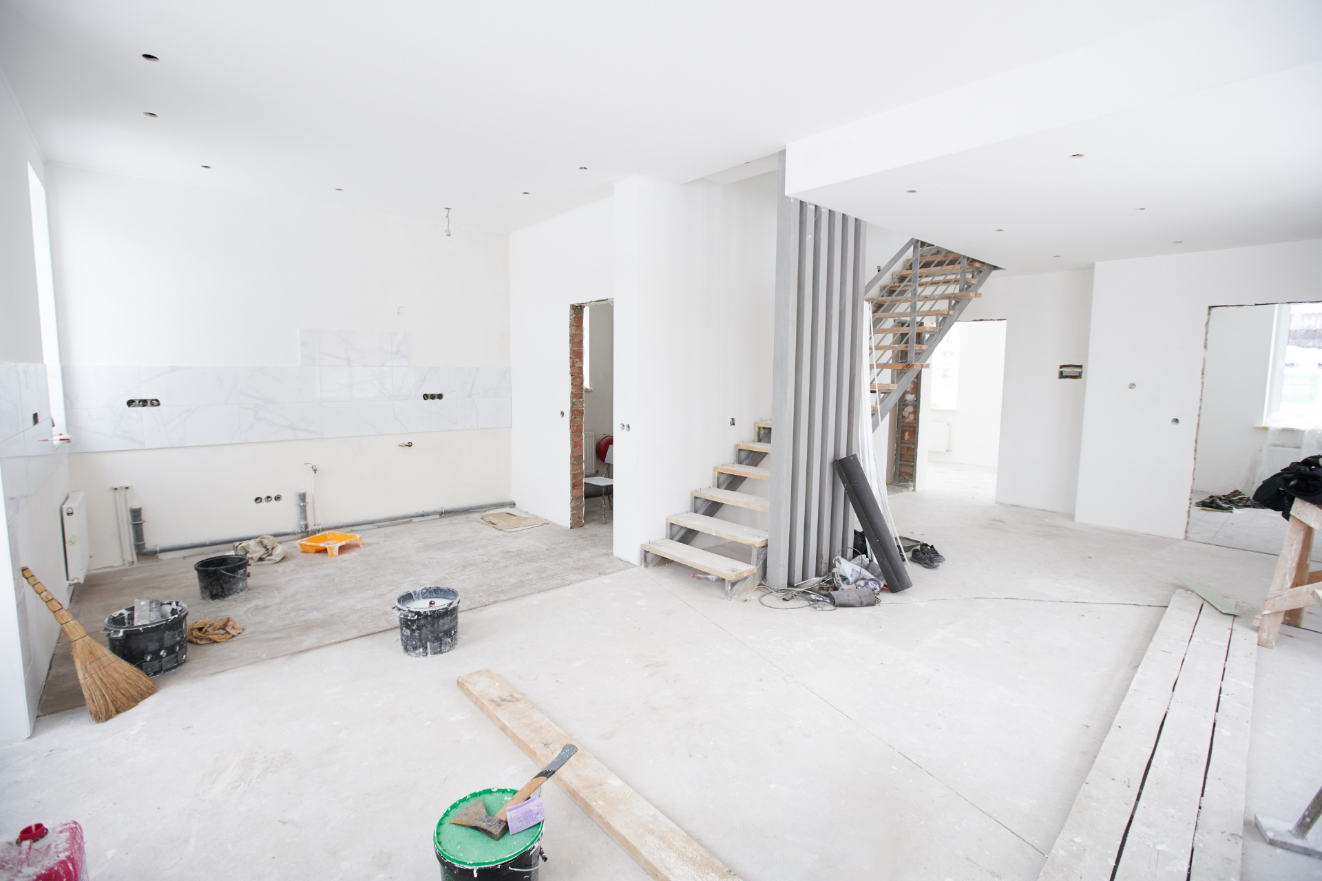 Bright unfinished interior room with white walls, staircase, tools, and construction materials scattered on the floor