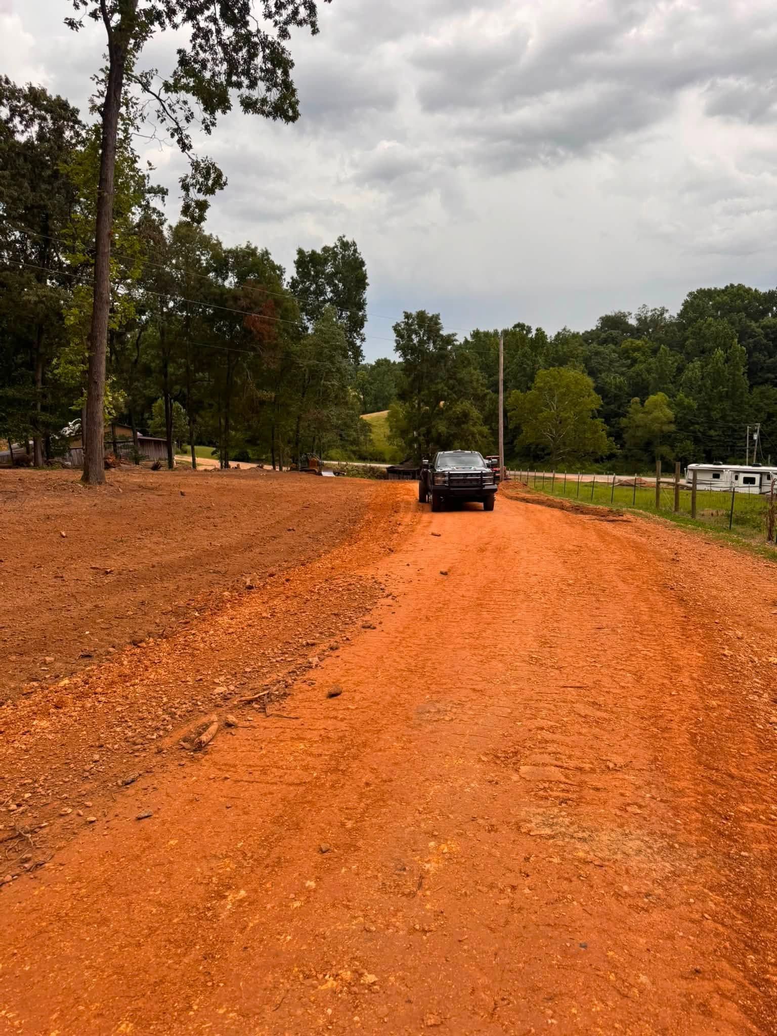 A car driving on a red dirt road beside trees under a cloudy sky.