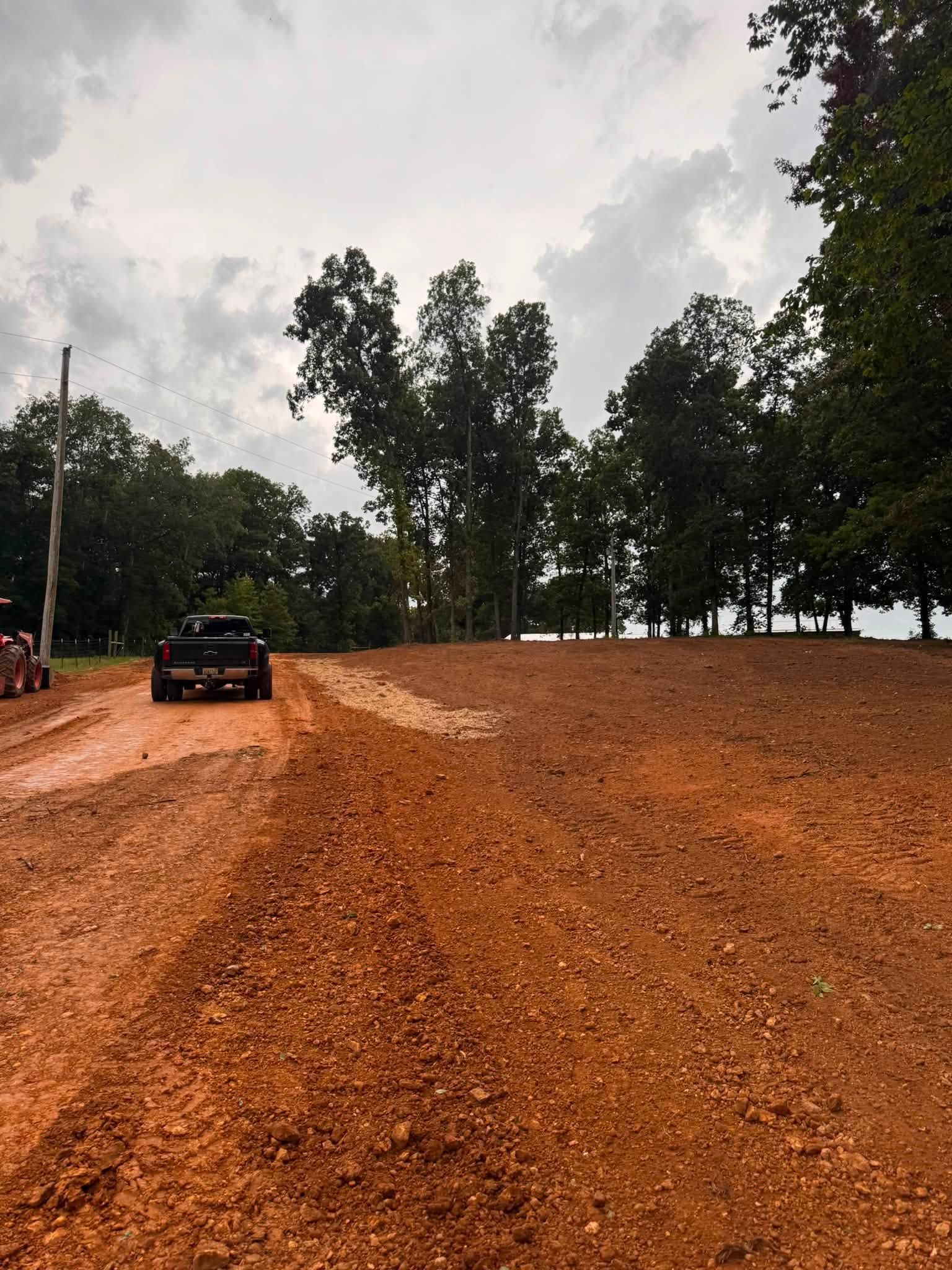 Red dirt road curving through a wooded area, with a black car parked on the left under a cloudy sky.