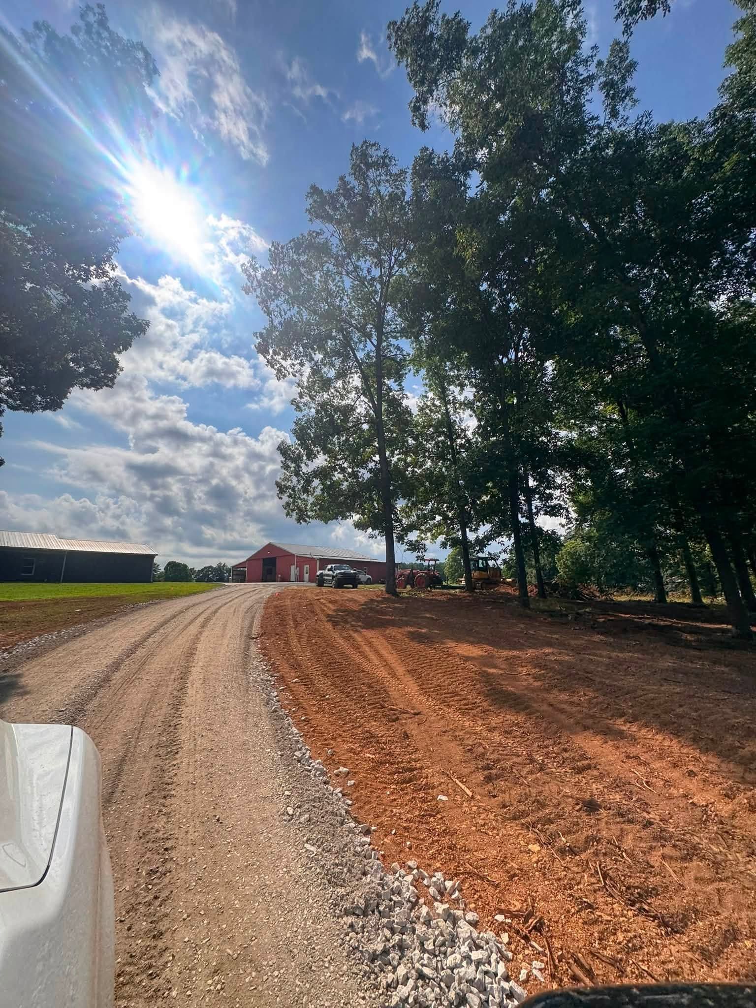 Gravel driveway curving past trees toward a red barn under a bright blue sky with clouds