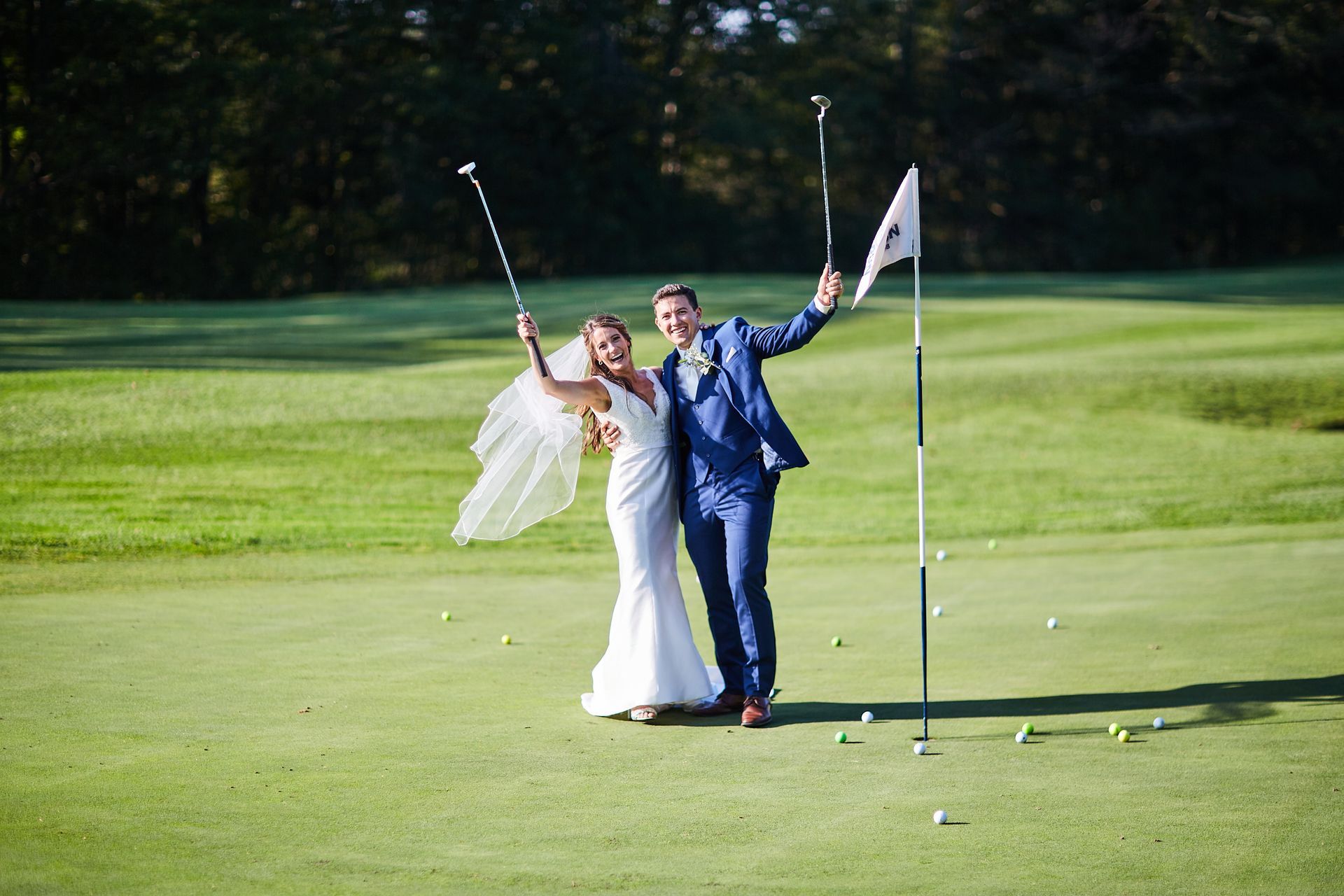 A bride and groom are standing on a golf course holding golf clubs and a flag.