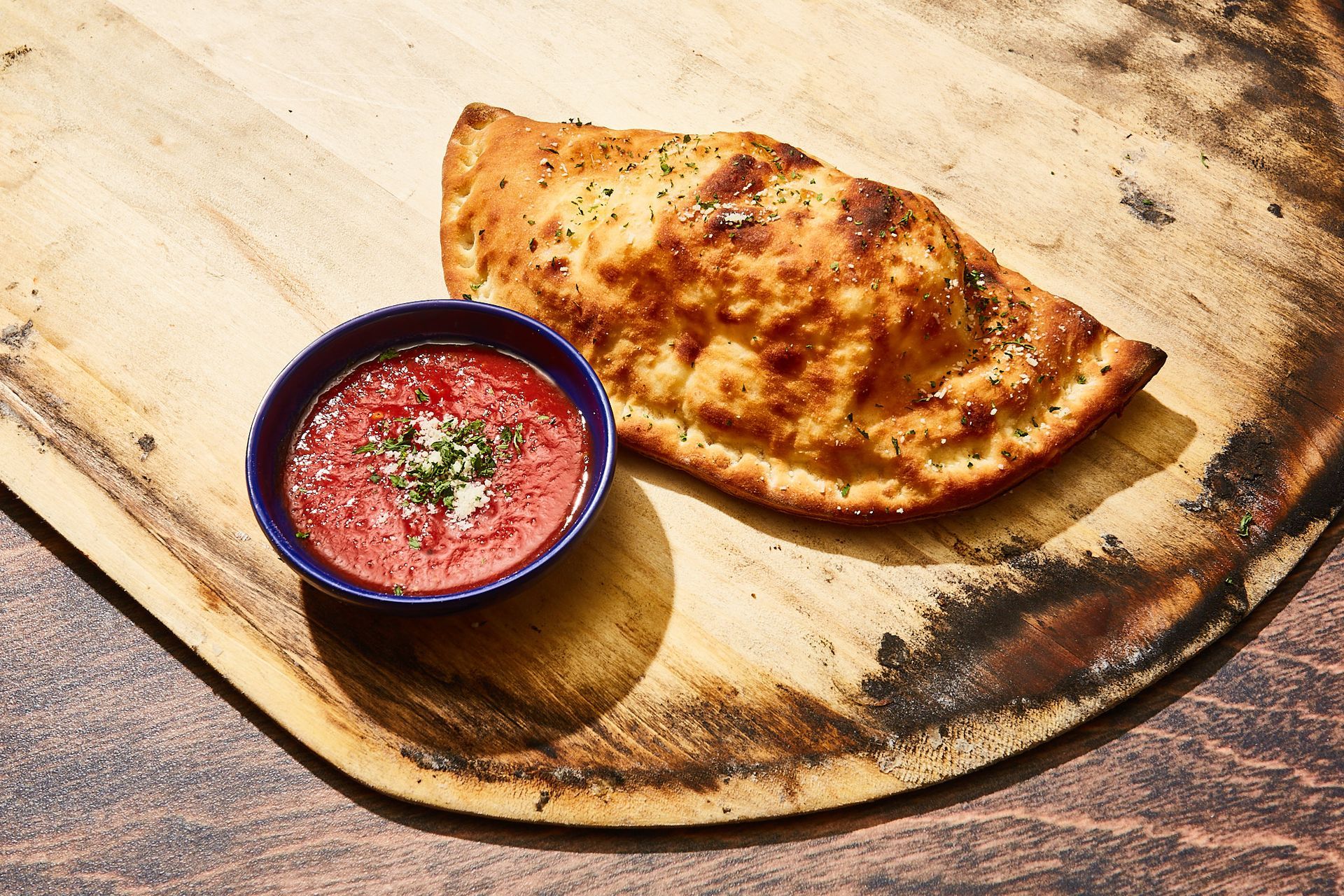 A calzone is sitting on a wooden cutting board next to a bowl of tomato sauce.