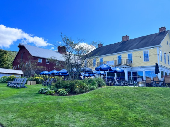 A large yellow building with blue umbrellas in front of it and a red barn in the background.
