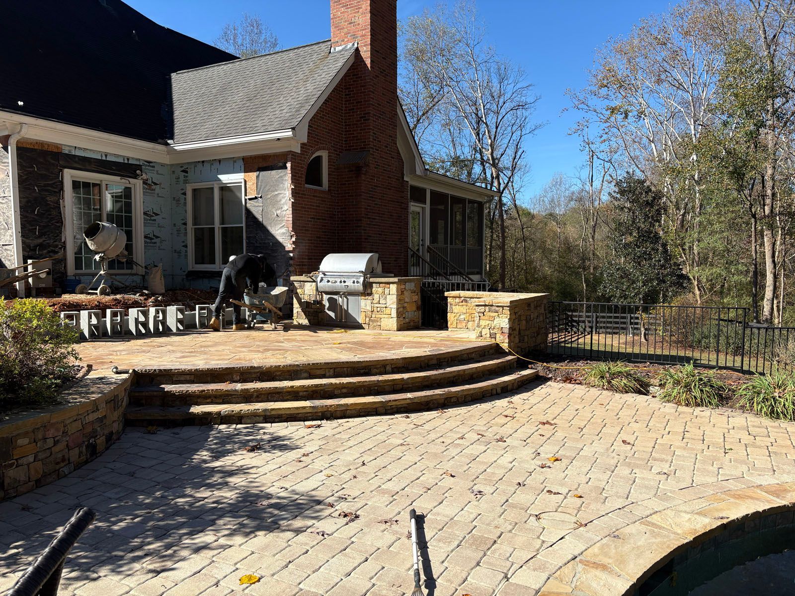 Stone patio with curved steps, brick house, outdoor grill, and wooded yard in sunlight