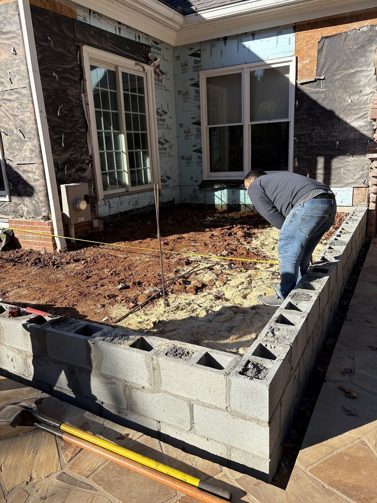 Worker bending over a raised cinder block patio wall during outdoor construction beside a house