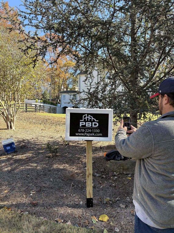 Person aiming a handgun at a black sign reading “PBO” in a wooded outdoor range.
