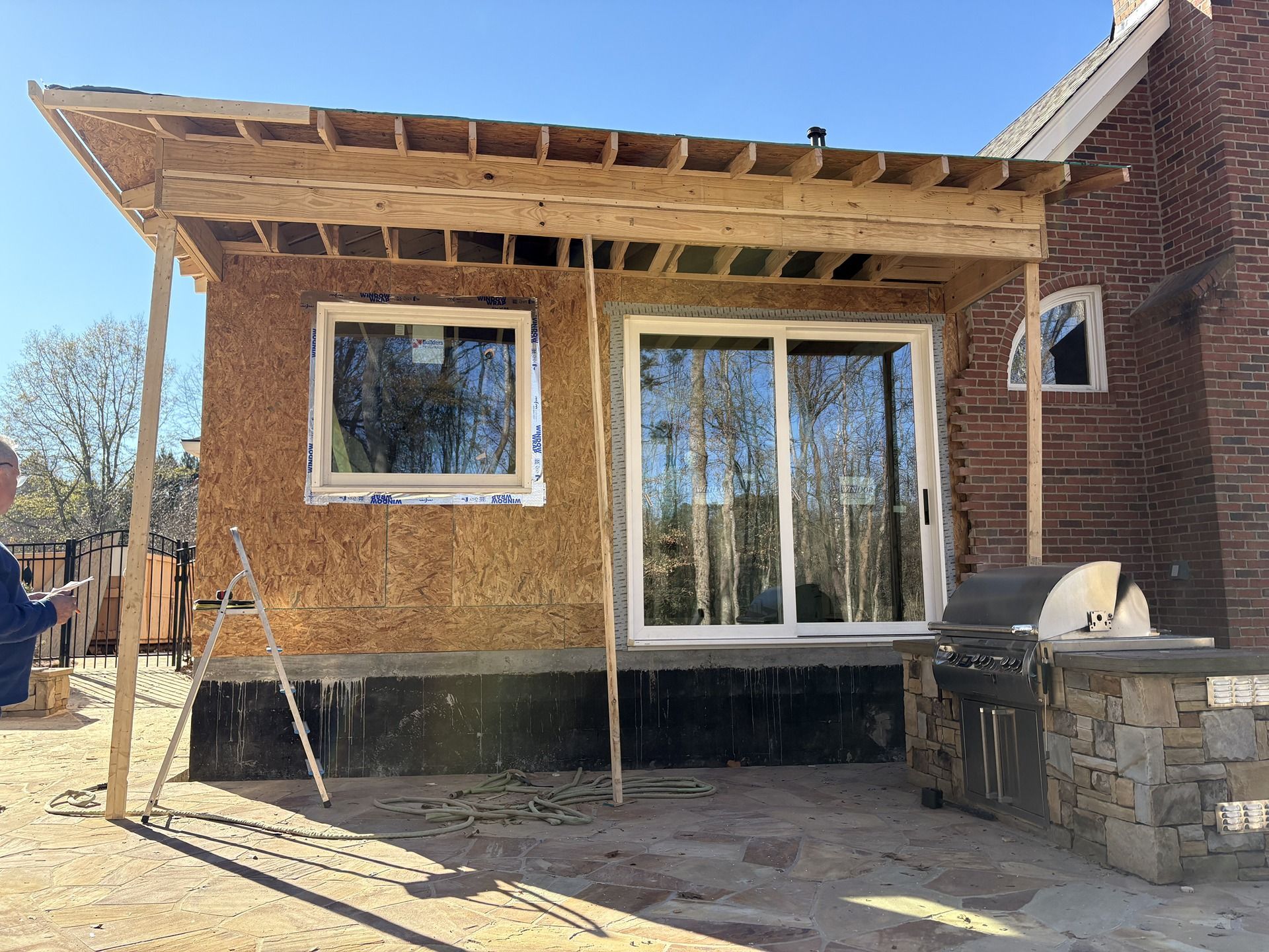 Backyard patio under construction with exposed framing, large windows, and a grill beside a brick house