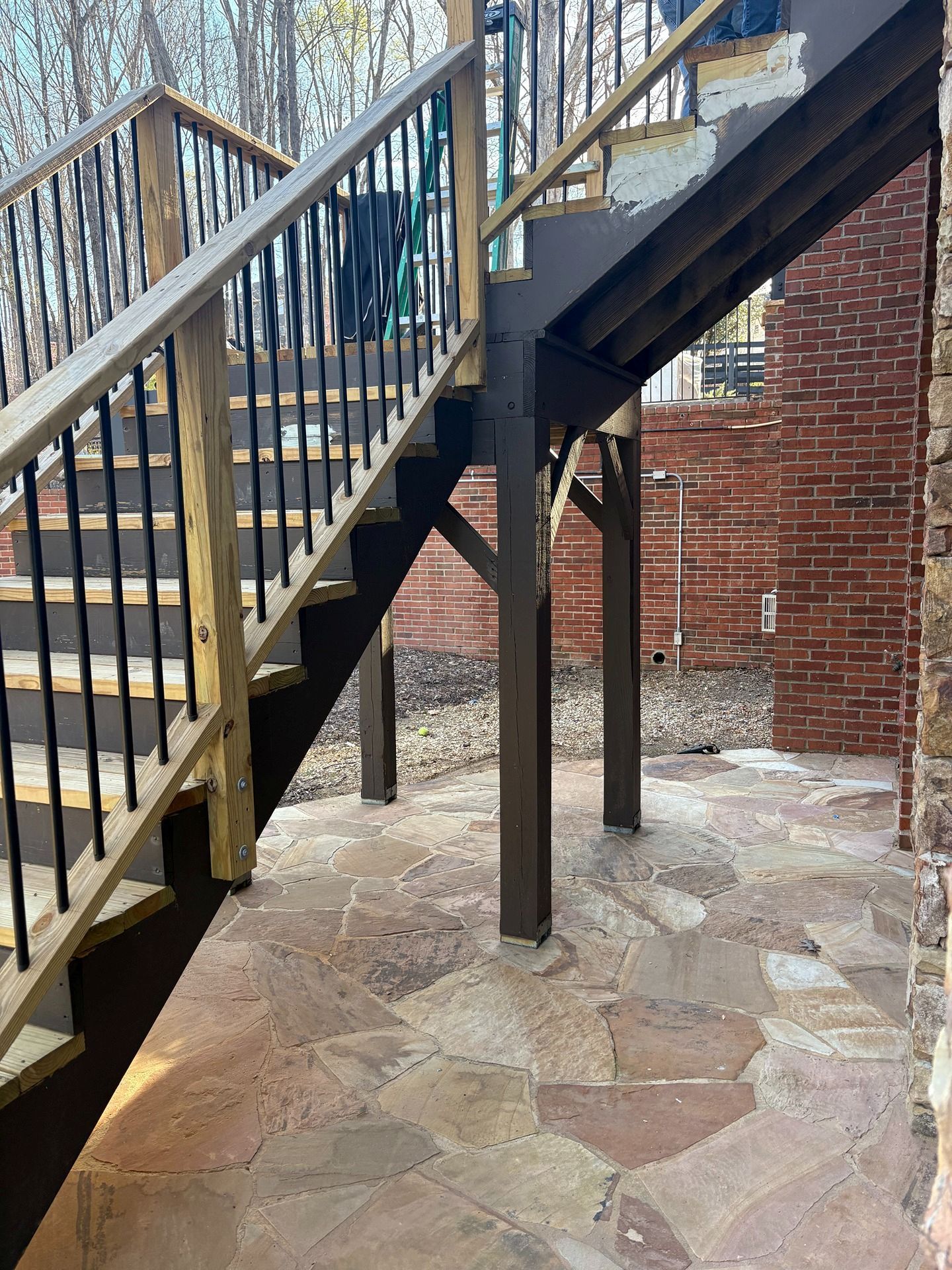 Outdoor staircase with wooden railings and support posts over a stone patio beside a brick wall.