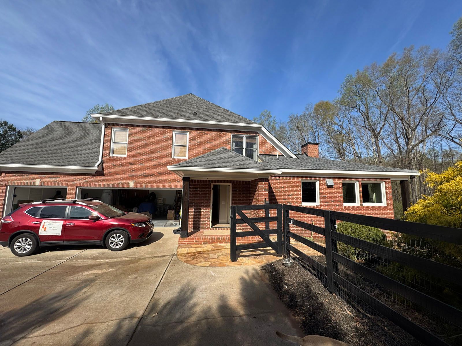 Brick house with a red SUV in the driveway, front porch, and a wooden fence under a blue sky.