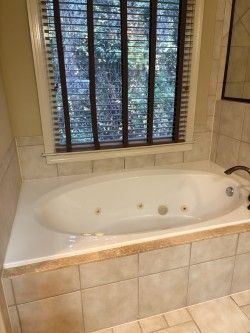 Bathroom with a white jetted bathtub set into beige tile beneath a window with blinds.