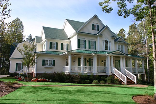Large white Victorian house with green shutters, wraparound porch, and lawn under a blue sky