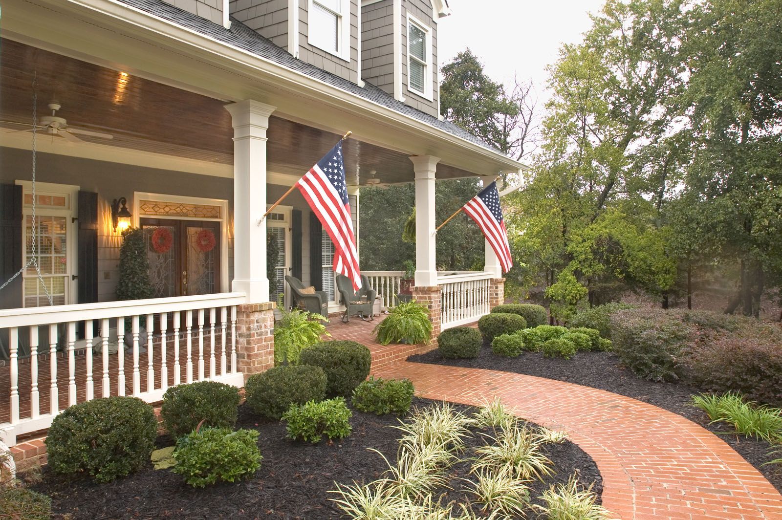 Front porch of a house with American flags, white columns, and a curved brick walkway through manicured shrubs
