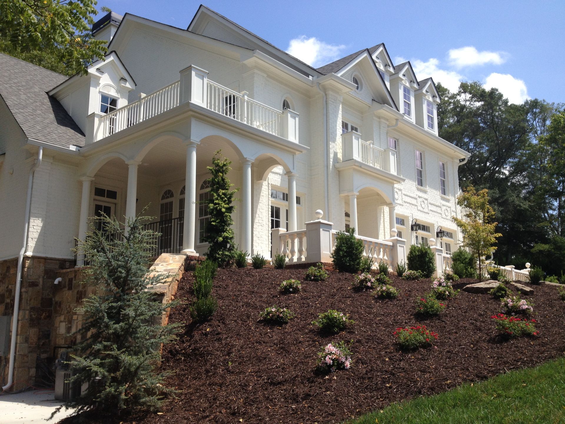 Large white house with balconies and porch on a landscaped hillside under a blue sky