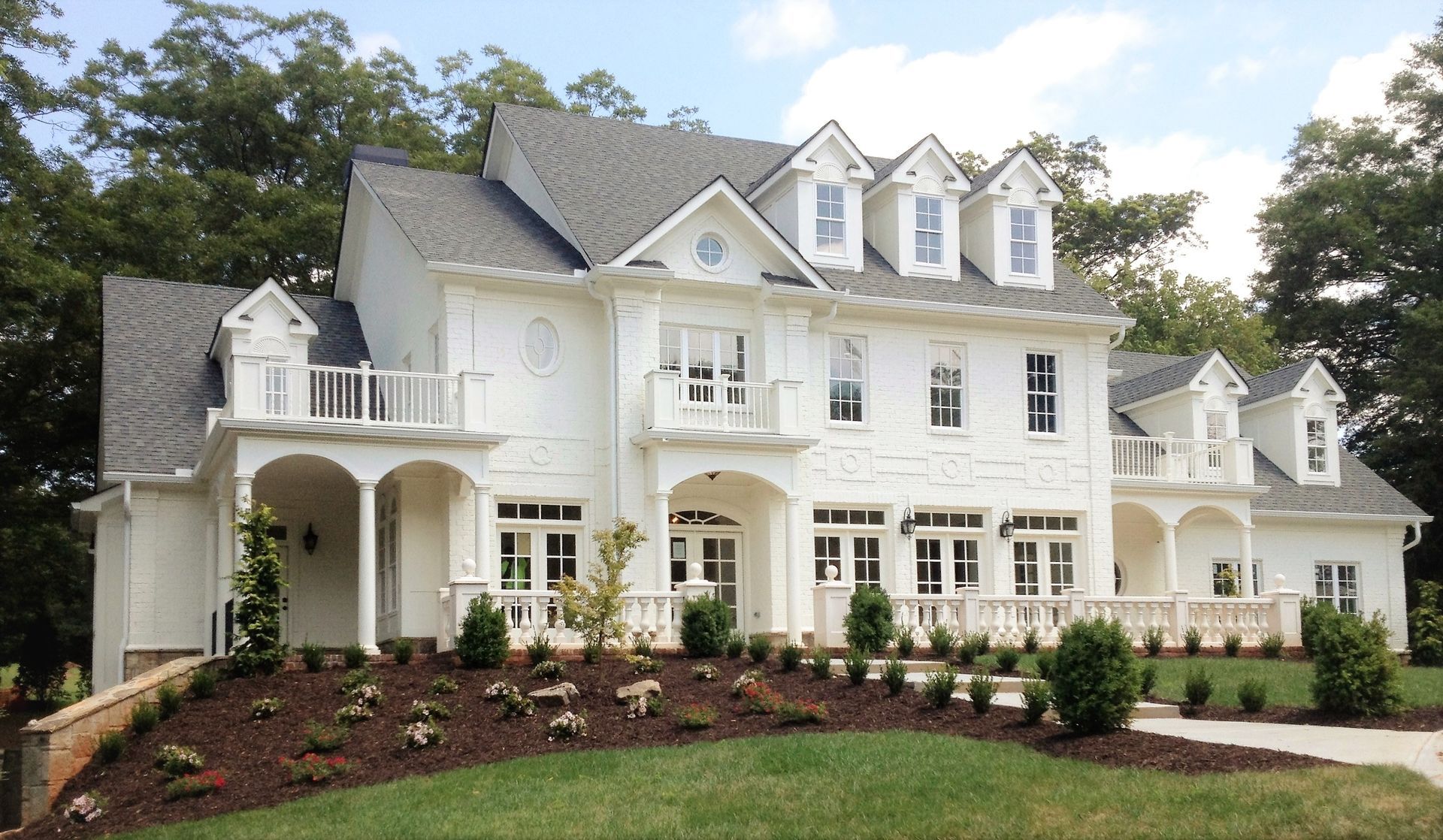 Large white two-story house with gray roof, balconies, and landscaped front yard