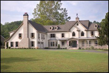 Large white country house with a porch and chimneys, set on a grassy lawn with trees behind it