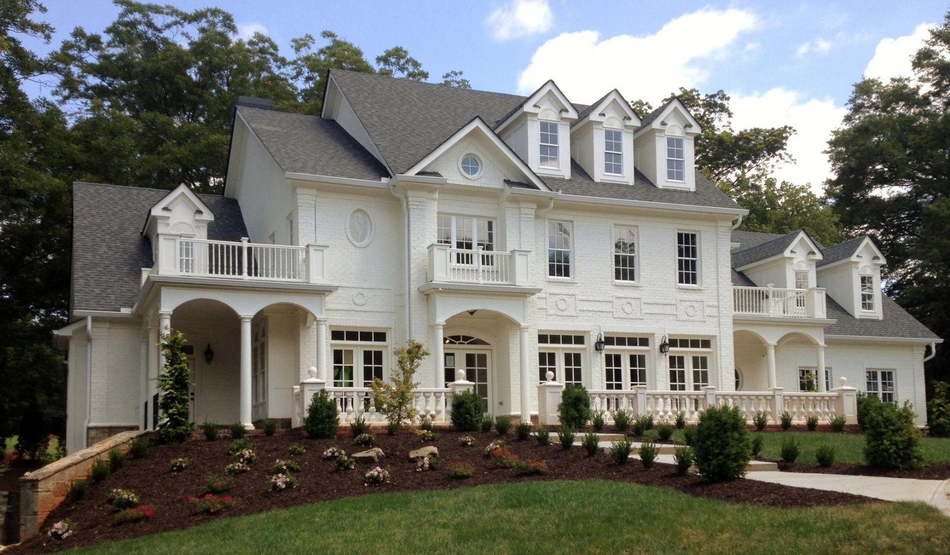 Large white suburban house with dormers, balconies, and landscaped front yard on a sunny day