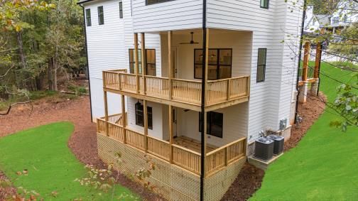 White two-story house with wooden wraparound deck and staircase, surrounded by trees and green lawn