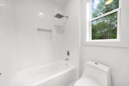 Bright white bathroom with bathtub, showerhead, toilet, and window showing trees outside.