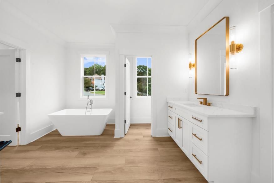 Bright modern bathroom with freestanding tub, white vanity, gold mirror, and wood floors.