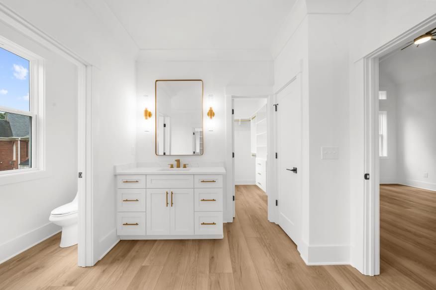 Bright modern bathroom with white vanity, gold mirror and fixtures, wood floor, and open door to another room