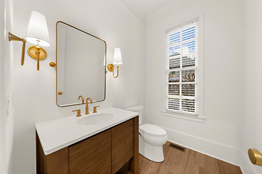 Bright white bathroom with wood vanity, gold fixtures, mirror, toilet, and window blinds.