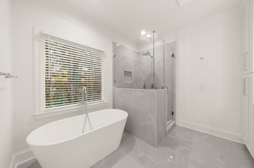 Bright white modern bathroom with freestanding tub, glass shower, and window blinds