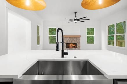 Modern white kitchen with large island, stainless sink, black faucet, and brick fireplace view.