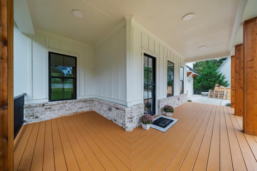 Covered front porch with tan decking, white siding, and black-trimmed windows, leading to an outdoor seating area.