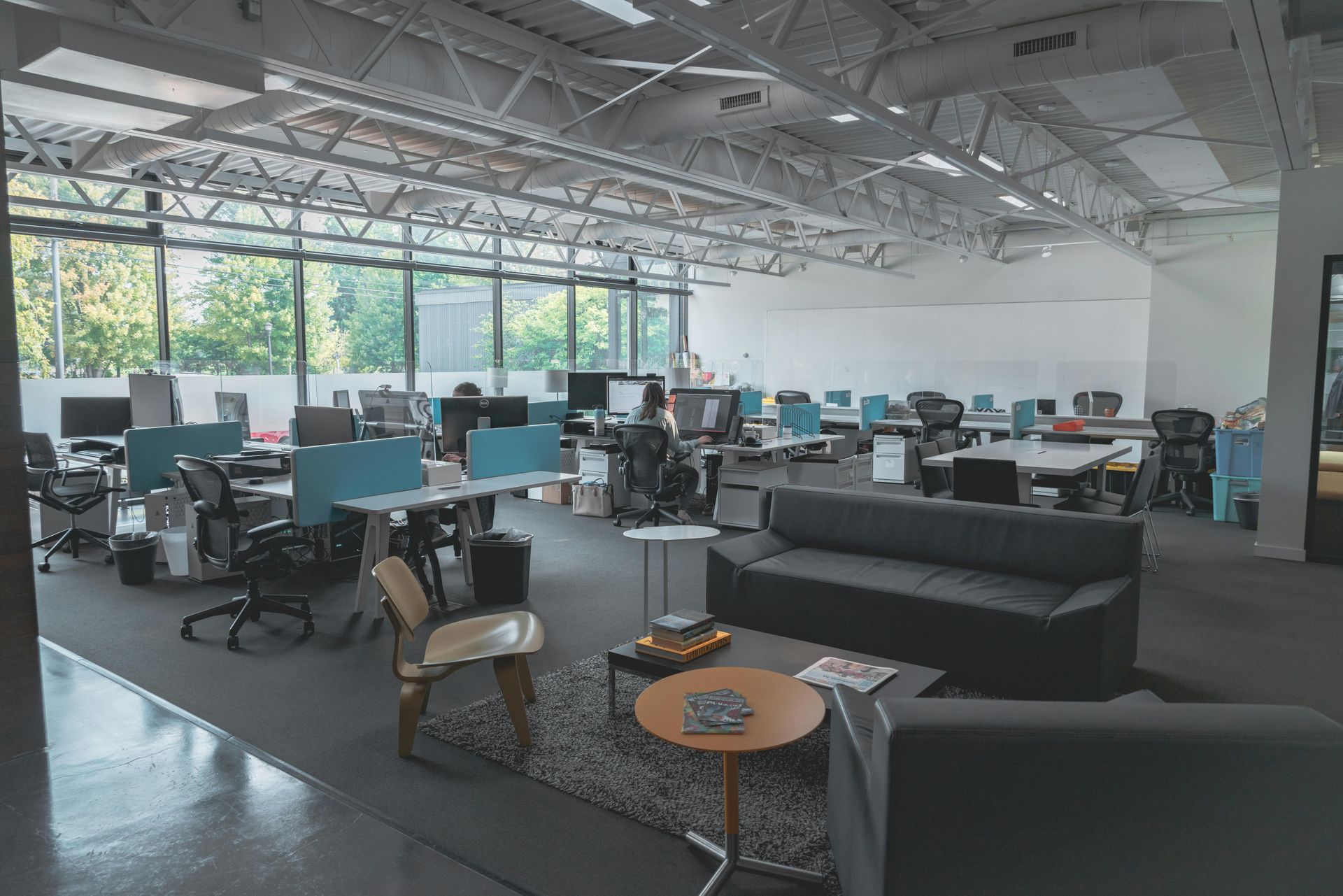 Open-plan office with rows of desks, computers, black sofas, and large windows letting in daylight