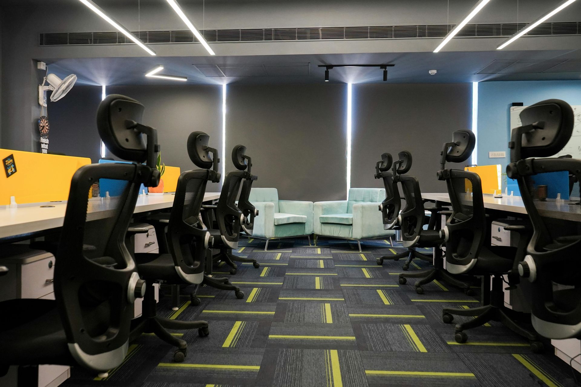 Modern office with rows of black swivel chairs and desks facing a central lounge area under bright strip lights