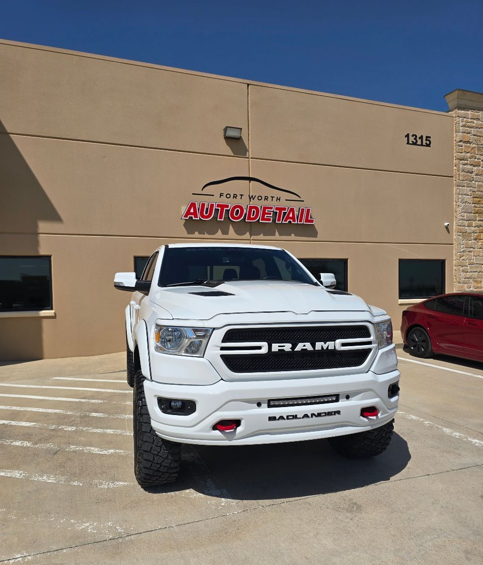 White Ram truck parked in front of an auto repair shop with red signage and a red car visible on the right.