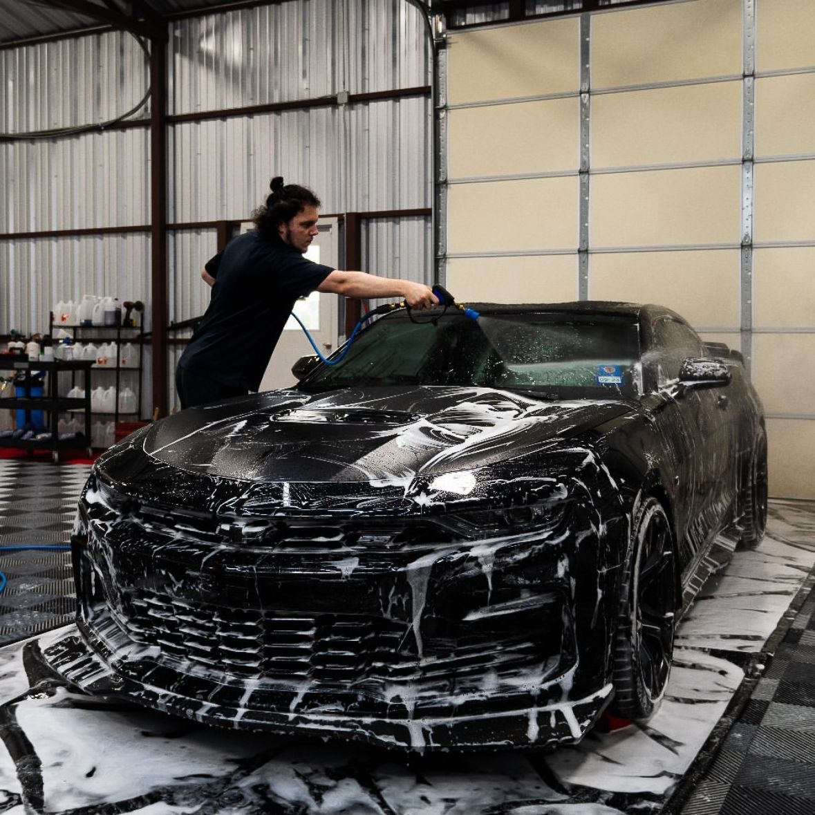 Person washing a black sports car in a garage, covered in soap foam