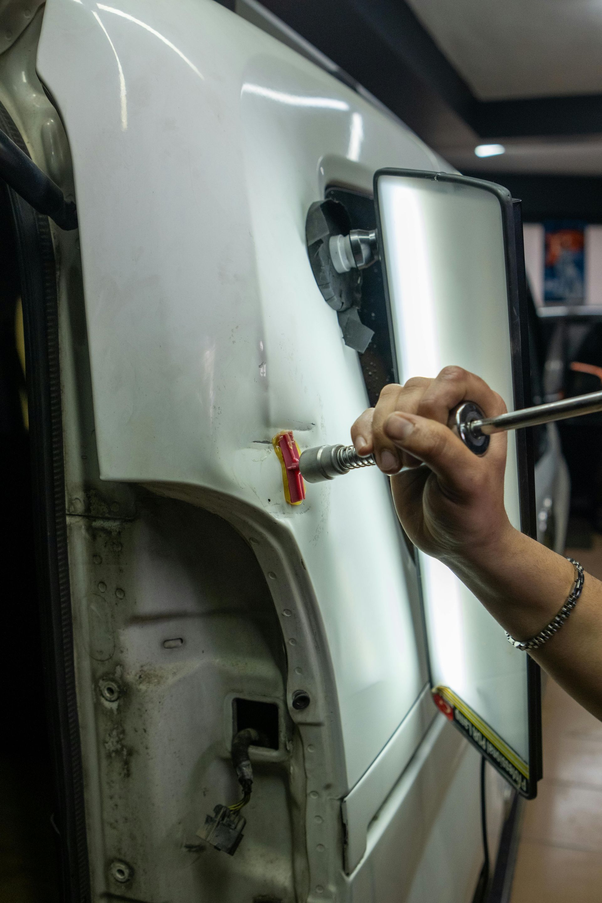A technician uses a glue pulling tool to remove a dent from a white vehicle panel, guided by a reflective light board.